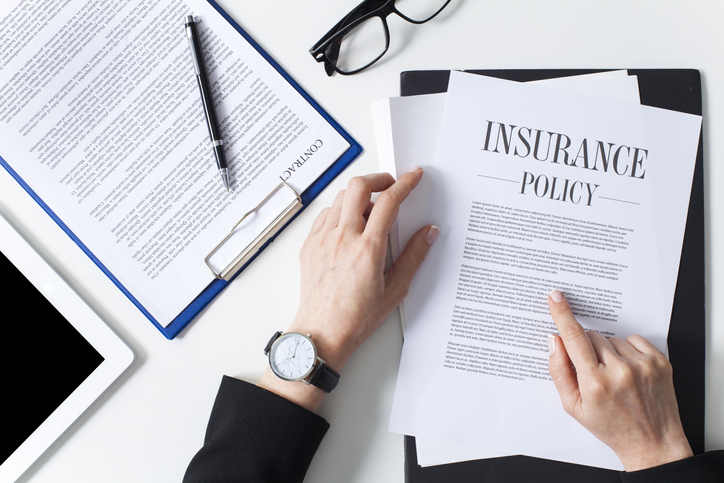 a desk with contracts, a pen, a pair of glases and a person pointing at the top page of an insurance policy.