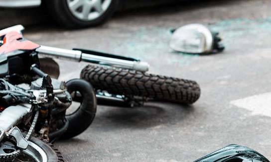 a motorcycle lays on its side after having a motor vehicle accident. Glass is shattered all over the ground and a helmet is laying in the distance near the wheels of the car that hit them. 