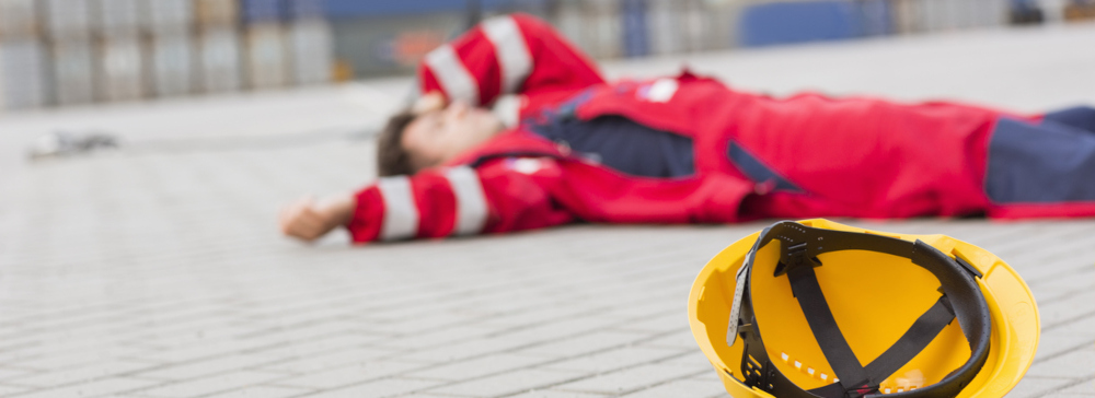 Construction worker in a red jumpsuit and yellow hardhat lying injured on concrete ground