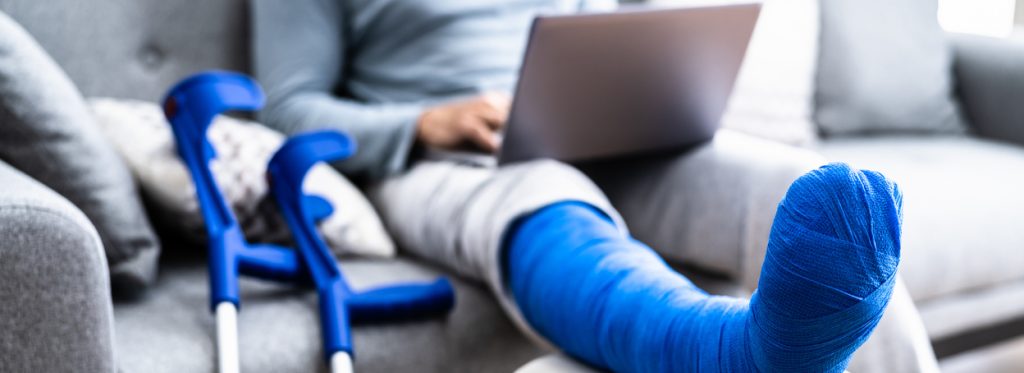 An injured man on the couch with a laptop and a blue cast over his broken leg