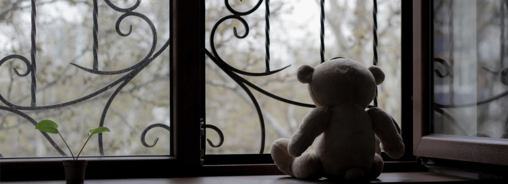 A teddy bear doll sitting on a dark windowsill looking out at a dreary day