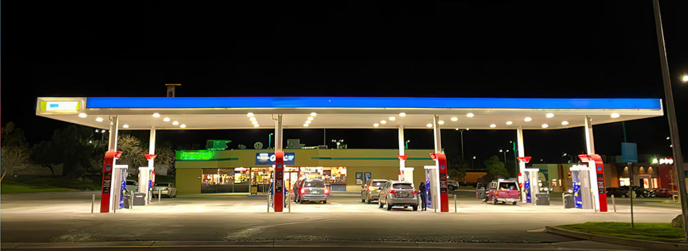 A brightly lit gas station at night, with vehicles refueling and a convenience store visible in the background.